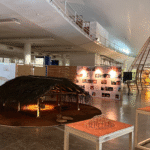 Foto mostra terceiro andar do pavilhão da Oca, no parque do Ibirapuera, em São Paulo, na 14ª Bienal Internacional de Arquitetura, em 2025, onde se vêem uma construção indígena, em primeiro plano, forrada de palha e, ao fundo, uma estrutura construída com Bambu, além de painéis fotográficos.
