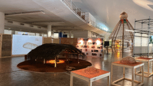 Foto mostra terceiro andar do pavilhão da Oca, no parque do Ibirapuera, em São Paulo, na 14ª Bienal Internacional de Arquitetura, em 2025, onde se vêem uma construção indígena, em primeiro plano, forrada de palha e, ao fundo, uma estrutura construída com Bambu, além de painéis fotográficos.