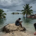 Imagem gerada por inteligência artificial mostra tela de celular em primeiro plano, no canto inferior direito, com homem sentado sobre um banco de areia, em segundo plano, com copas de árvores e telhados de casas acima do nível da água em local inundado pelo mar.
