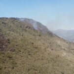 Fotografia aérea da encosta de uma montanha na Serra do Cipó, em Minas Gerais. A imagem mostra fumaça branca subindo da vegetação seca em direção ao céu azul e grandes áreas de solo e rochas já carbonizadas, de cor preta, no lado esquerdo da encosta.