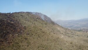 Fotografia aérea da encosta de uma montanha na Serra do Cipó, em Minas Gerais. A imagem mostra fumaça branca subindo da vegetação seca em direção ao céu azul e grandes áreas de solo e rochas já carbonizadas, de cor preta, no lado esquerdo da encosta.
