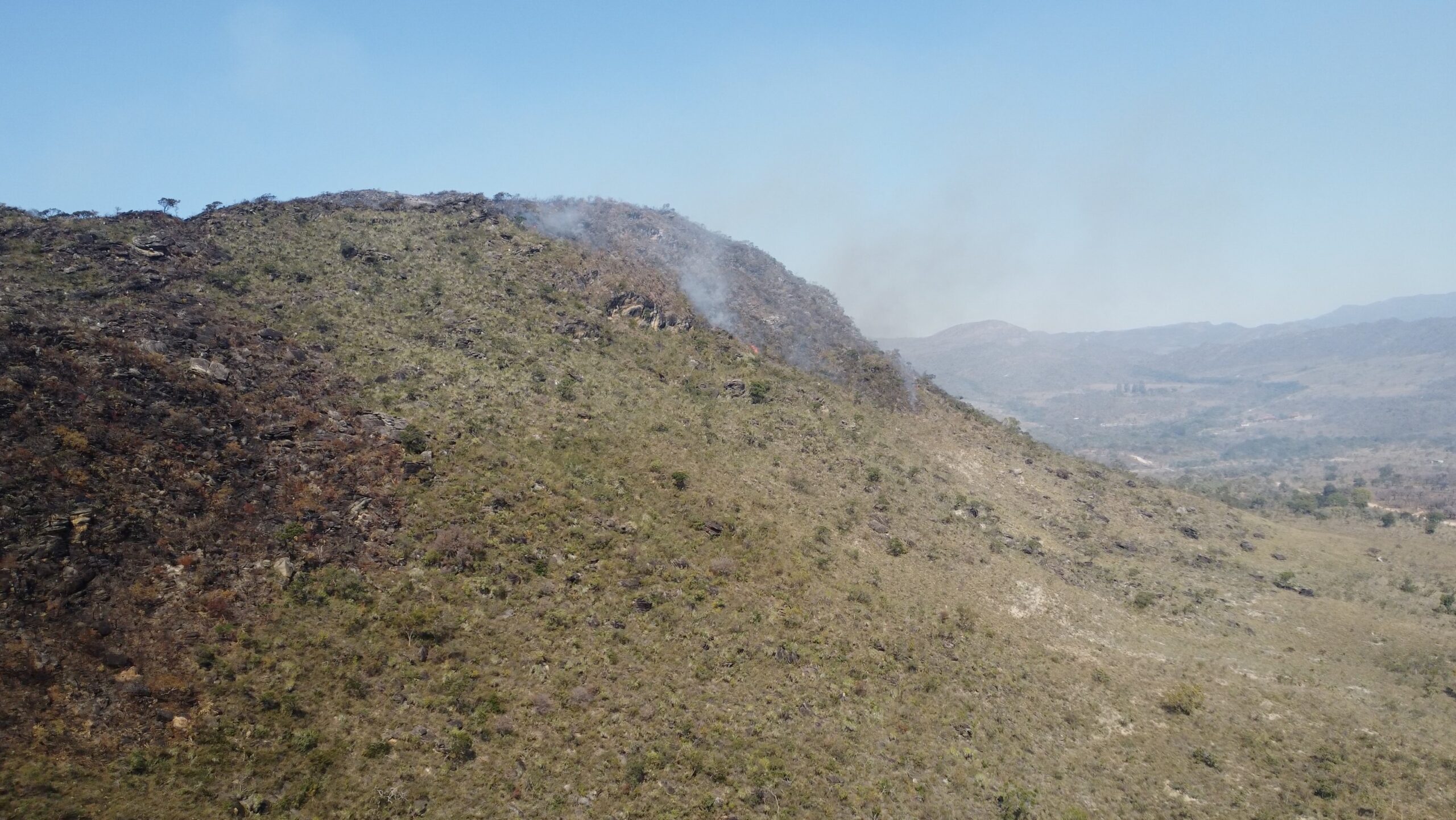 Fotografia aérea da encosta de uma montanha na Serra do Cipó, em Minas Gerais. A imagem mostra fumaça branca subindo da vegetação seca em direção ao céu azul e grandes áreas de solo e rochas já carbonizadas, de cor preta, no lado esquerdo da encosta.