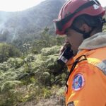 No canto direito da foto, um homem com uniforme de combate ao fogo, macacão e capacete do corpo de Bombeiros de Minas Gerais, fala ao rádio. Ao fundo uma encosta da Serra do Brigadeiro, de mata verde, são vistas colunas de fumaça saindo de dentro da vegetação.