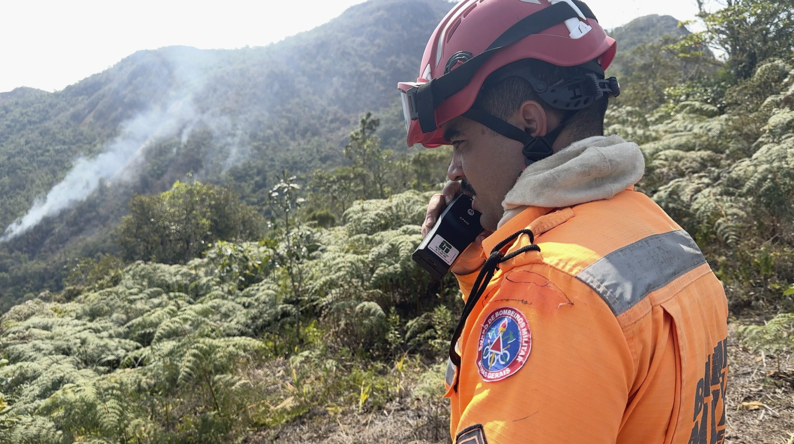 No canto direito da foto, um homem com uniforme de combate ao fogo, macacão e capacete do corpo de Bombeiros de Minas Gerais, fala ao rádio. Ao fundo uma encosta da Serra do Brigadeiro, de mata verde, são vistas colunas de fumaça saindo de dentro da vegetação.