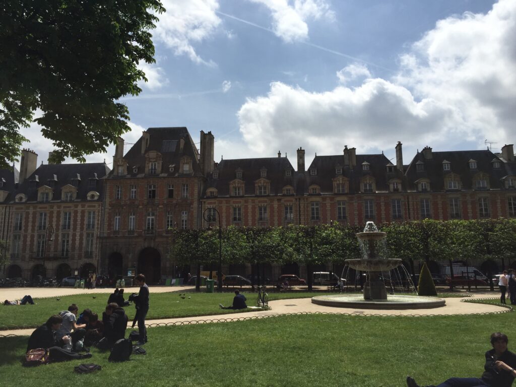 Fotografia de arquivo da Praça de Vosges em Paris, França, mostrando o gramado central com pessoas relaxando em um piquenique, uma fonte de água ativa e edifícios históricos de tijolos vermelhos e pedra ao fundo sob um céu parcialmente nublado.