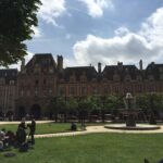 Fotografia de arquivo da Praça de Vosges em Paris, França, mostrando o gramado central com pessoas relaxando em um piquenique, uma fonte de água ativa e edifícios históricos de tijolos vermelhos e pedra ao fundo sob um céu parcialmente nublado.