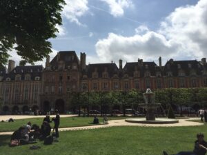 Fotografia de arquivo da Praça de Vosges em Paris, França, mostrando o gramado central com pessoas relaxando em um piquenique, uma fonte de água ativa e edifícios históricos de tijolos vermelhos e pedra ao fundo sob um céu parcialmente nublado.