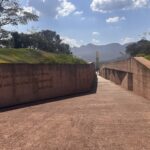 Fotografia de um caminho aberto no Memorial de Brumadinho durante o dia. O percurso é pavimentado e ladeado por extensos muros de concreto. No muro esquerdo, está cravada a citação: "O que a memória ama fica eterno. Te amo com a memória, imperecível", da poeta Adélia Prado. Ao fundo, vê-se um monumento vertical, árvores, montanhas e um céu azul com algumas nuvens.