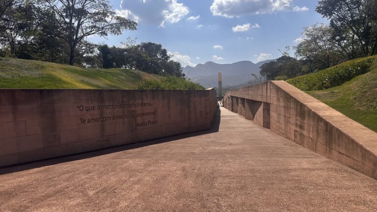 Fotografia de um caminho aberto no Memorial de Brumadinho durante o dia. O percurso é pavimentado e ladeado por extensos muros de concreto. No muro esquerdo, está cravada a citação: "O que a memória ama fica eterno. Te amo com a memória, imperecível", da poeta Adélia Prado. Ao fundo, vê-se um monumento vertical, árvores, montanhas e um céu azul com algumas nuvens.