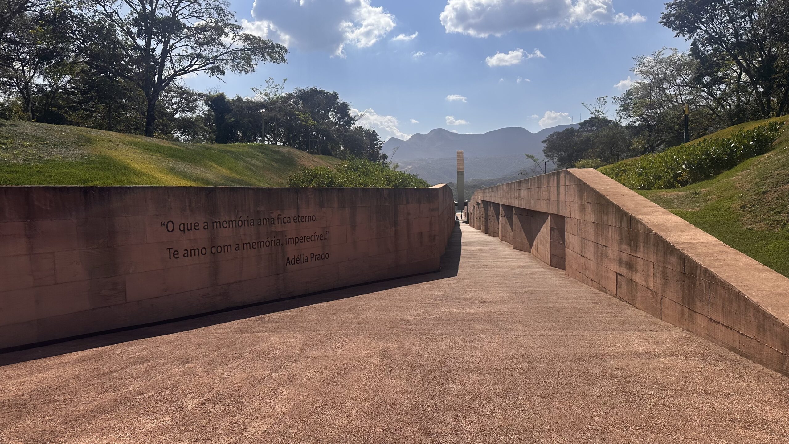 Fotografia de um caminho aberto no Memorial de Brumadinho durante o dia. O percurso é pavimentado e ladeado por extensos muros de concreto. No muro esquerdo, está cravada a citação: "O que a memória ama fica eterno. Te amo com a memória, imperecível", da poeta Adélia Prado. Ao fundo, vê-se um monumento vertical, árvores, montanhas e um céu azul com algumas nuvens.