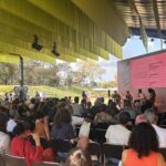 Fotografia de uma plateia numerosa sentada sob uma grande estrutura de lona amarela, assistindo a um painel com quatro palestrantes. Ao fundo, uma grande tela rosa exibe os dados da apresentação. A imagem ilustra a realização de debates sobre memória e meio ambiente no Memorial de Brumadinho.