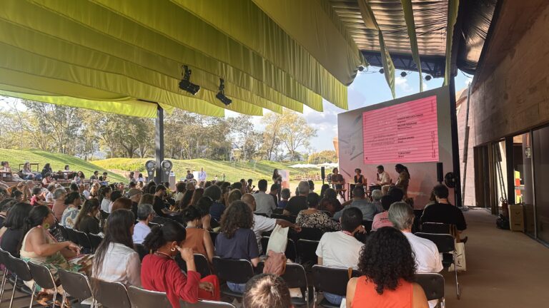 Fotografia de uma plateia numerosa sentada sob uma grande estrutura de lona amarela, assistindo a um painel com quatro palestrantes. Ao fundo, uma grande tela rosa exibe os dados da apresentação. A imagem ilustra a realização de debates sobre memória e meio ambiente no Memorial de Brumadinho.