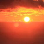 Sol brilhante no horizonte do oceano sob um céu vermelho intenso e nuvens escuras, imagem utilizada para representar o aquecimento global e as ondas de calor extremo.