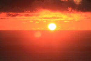 Sol brilhante no horizonte do oceano sob um céu vermelho intenso e nuvens escuras, imagem utilizada para representar o aquecimento global e as ondas de calor extremo.