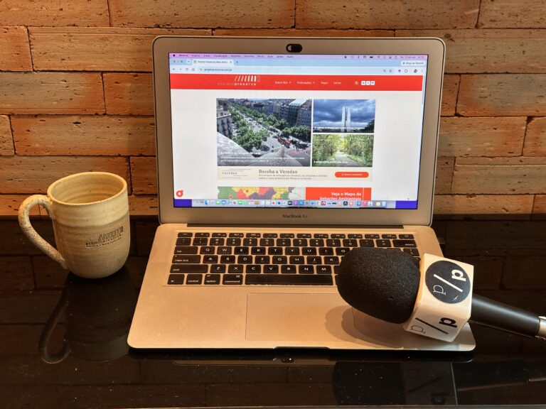 Fotografia de um ambiente de trabalho jornalístico, contendo um notebook aberto no site do Projeto Preserva, um microfone de reportagem e uma caneca, ilustrando a estrutura de produção da mídia independente.